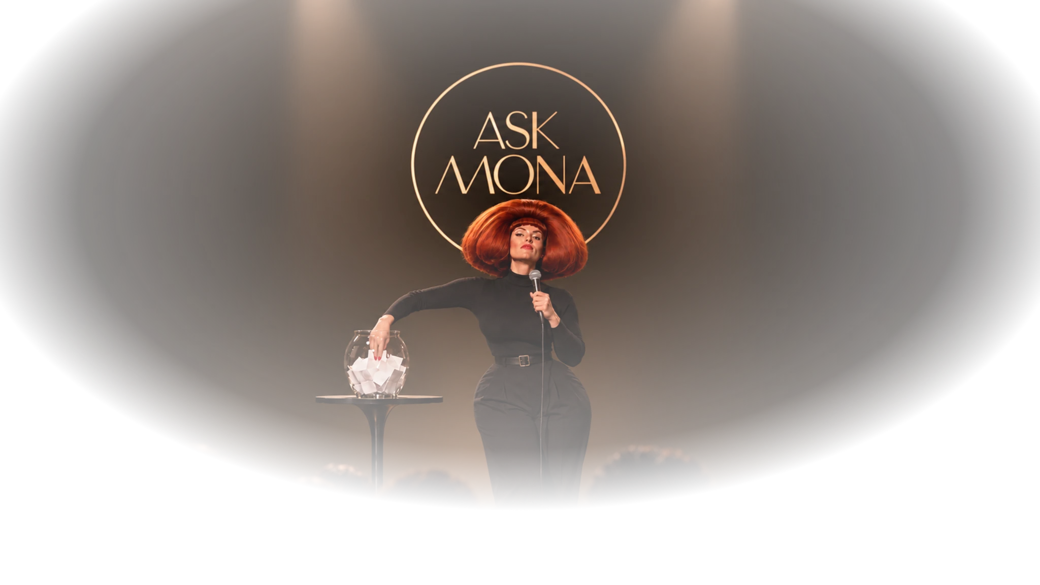 Mona Vex stands beneath the circular Ask Mona stage sign, holding a microphone and drawing a question from a glass bowl